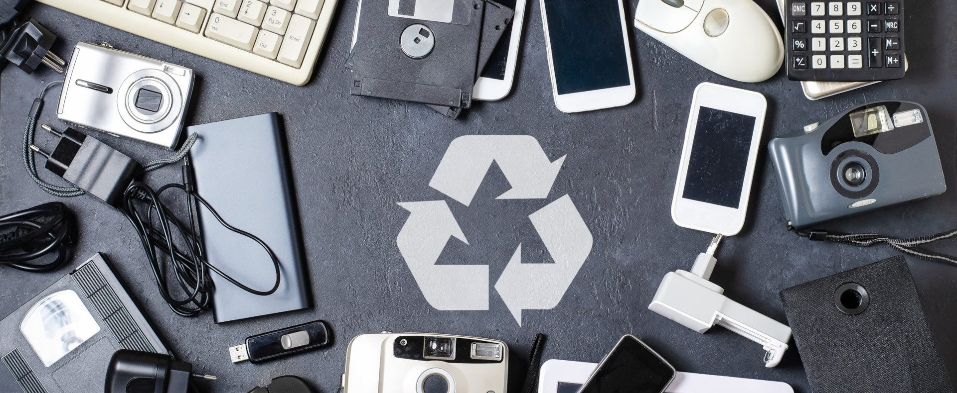broken phones, cameras, keyboards and more on a grey table with a white recycling logo in the centre